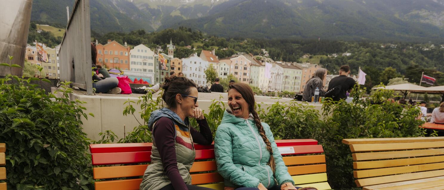 Two smiling women on a rainbow bench overlooking Innsbruck's old town and mountain panorama.