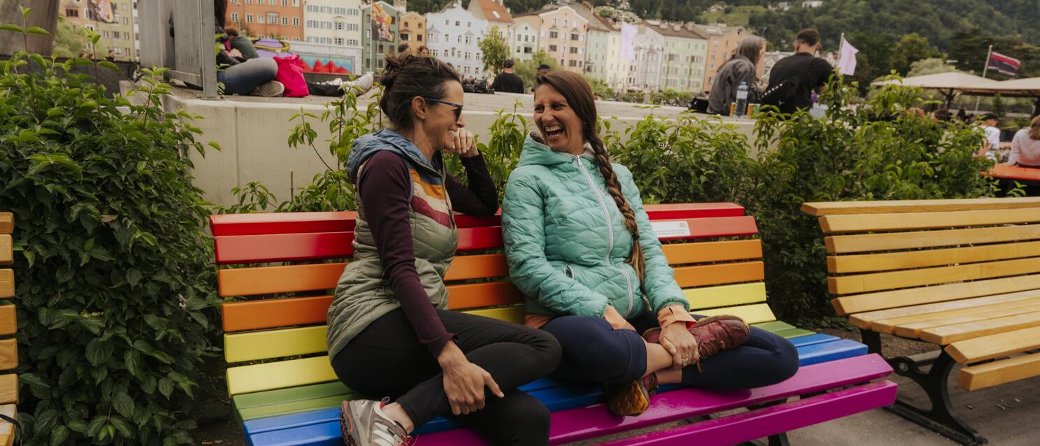 Two smiling women on a rainbow bench overlooking Innsbruck's old town and mountain panorama.