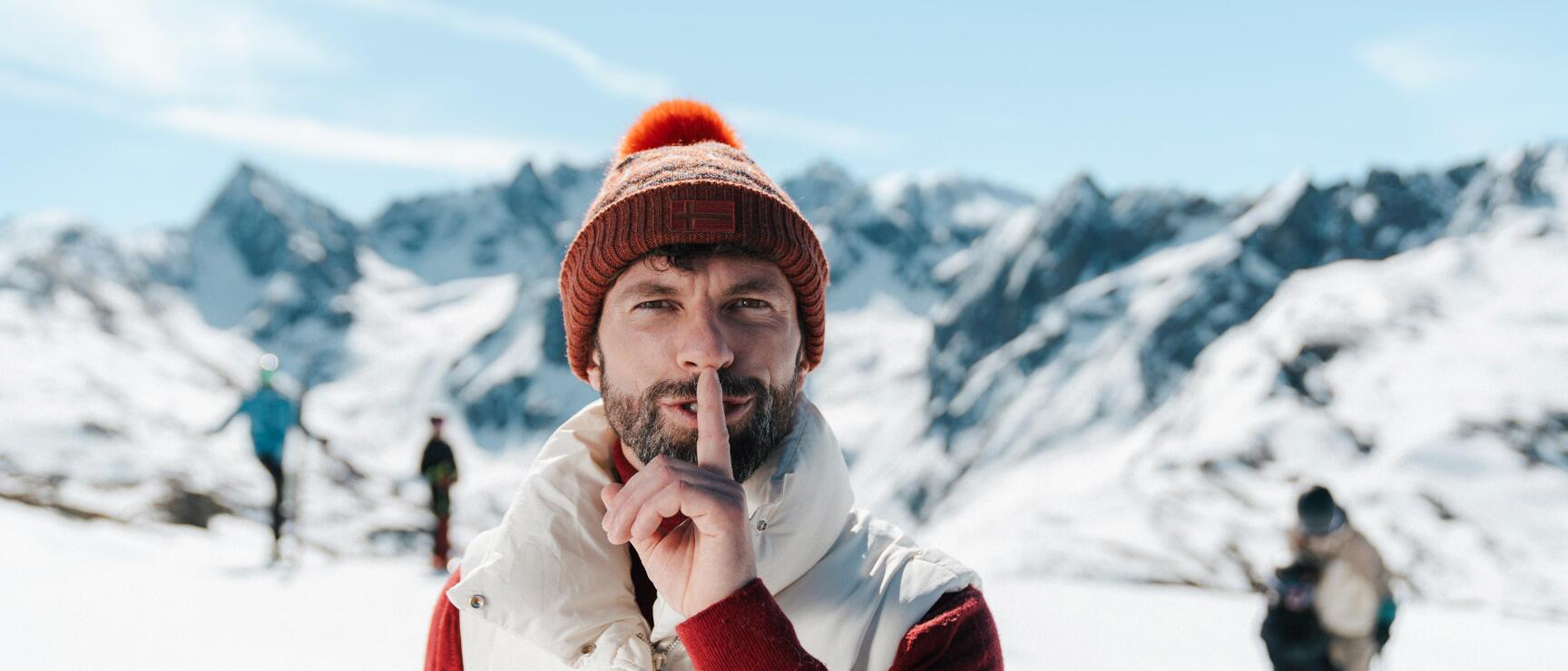 Man with orange-red wool hat holds finger to lips, snow-covered mountains and skiers in background.