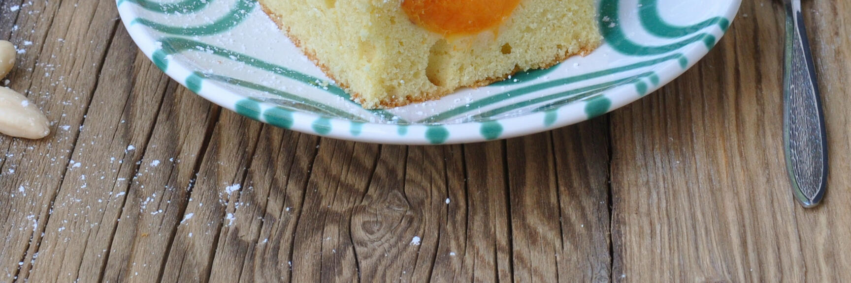 Slice of apricot cake with icing sugar on patterned plate, fresh apricots and mint on wooden table.