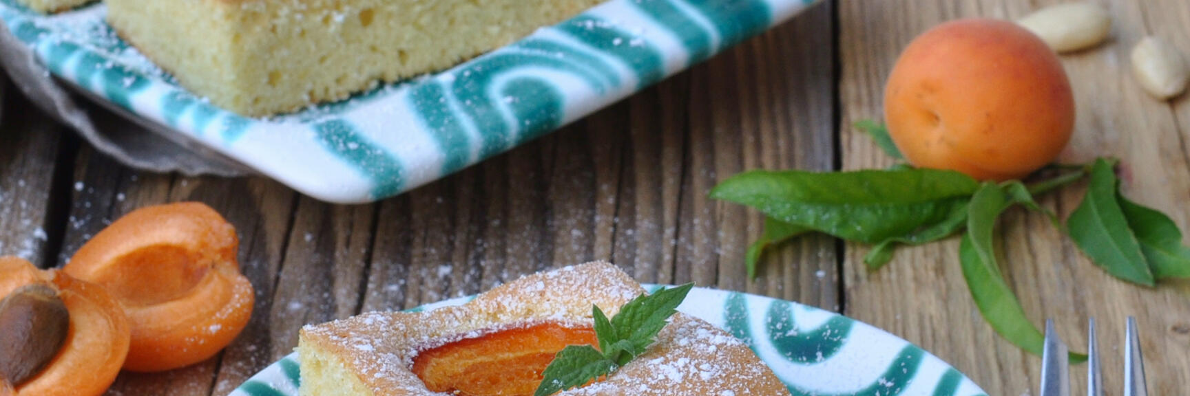 Slice of apricot cake with icing sugar on patterned plate, fresh apricots and mint on wooden table.