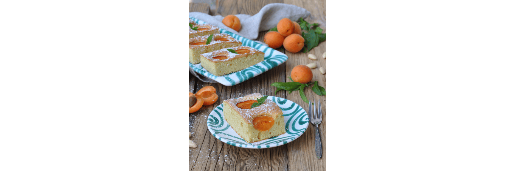Slice of apricot cake with icing sugar on patterned plate, fresh apricots and mint on wooden table.