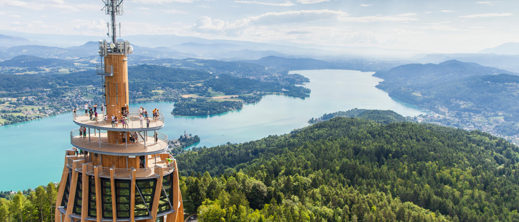 Holzaussichtsturm mit Besuchern auf der Plattform, Blick auf den Wörthersee und bewaldete Hügel, Kärnten.