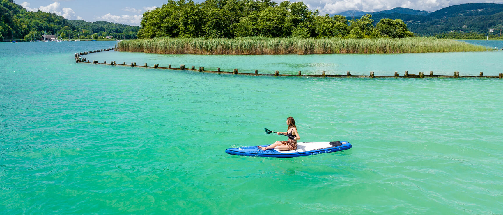 Frau auf SUP-Board paddelt auf türkisem See, Schilfinsel und Hügel im Hintergrund.