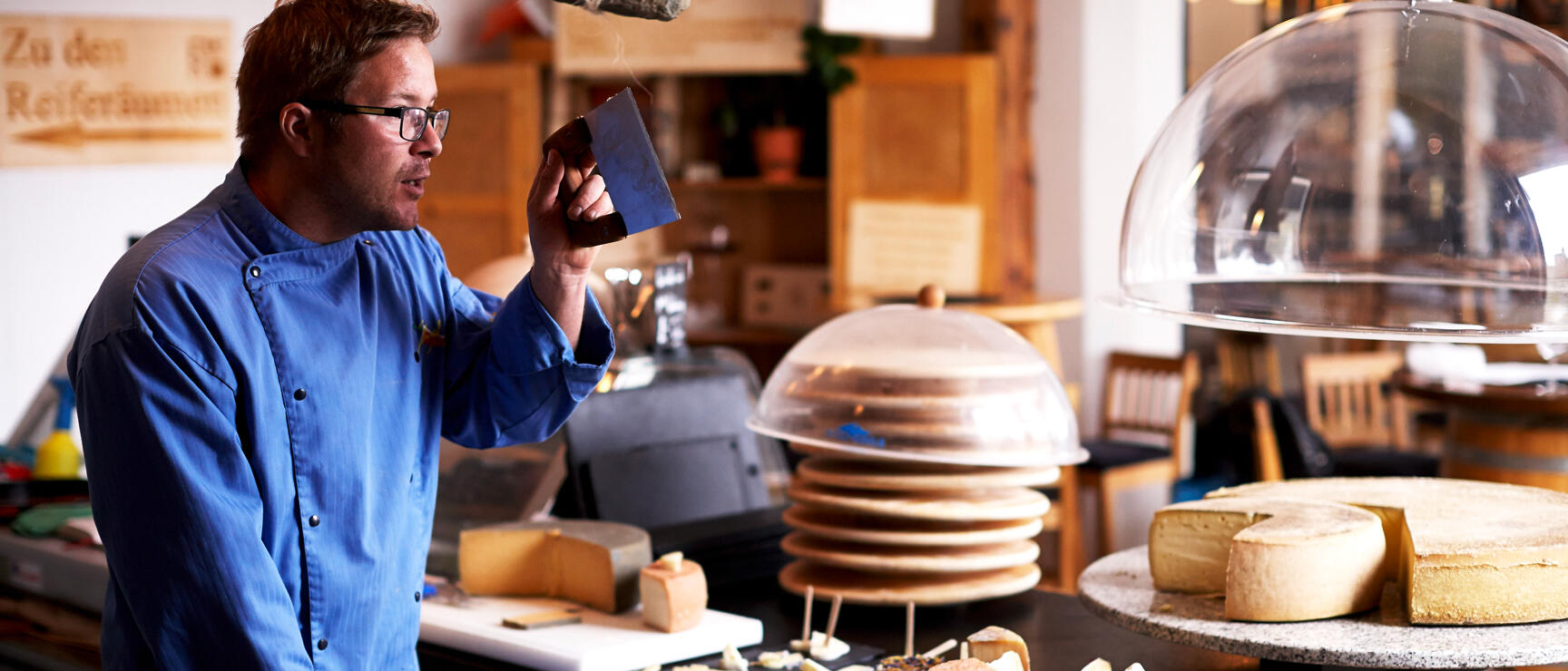 Man with glasses and blue shirt holding up piece of cheese in front of cheese counter with various cheese types.