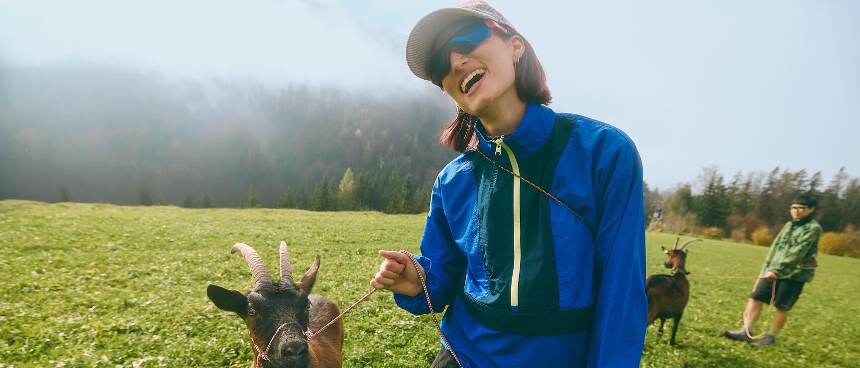 Laughing woman in blue jacket holding a brown goat on a leash on a green alpine meadow.