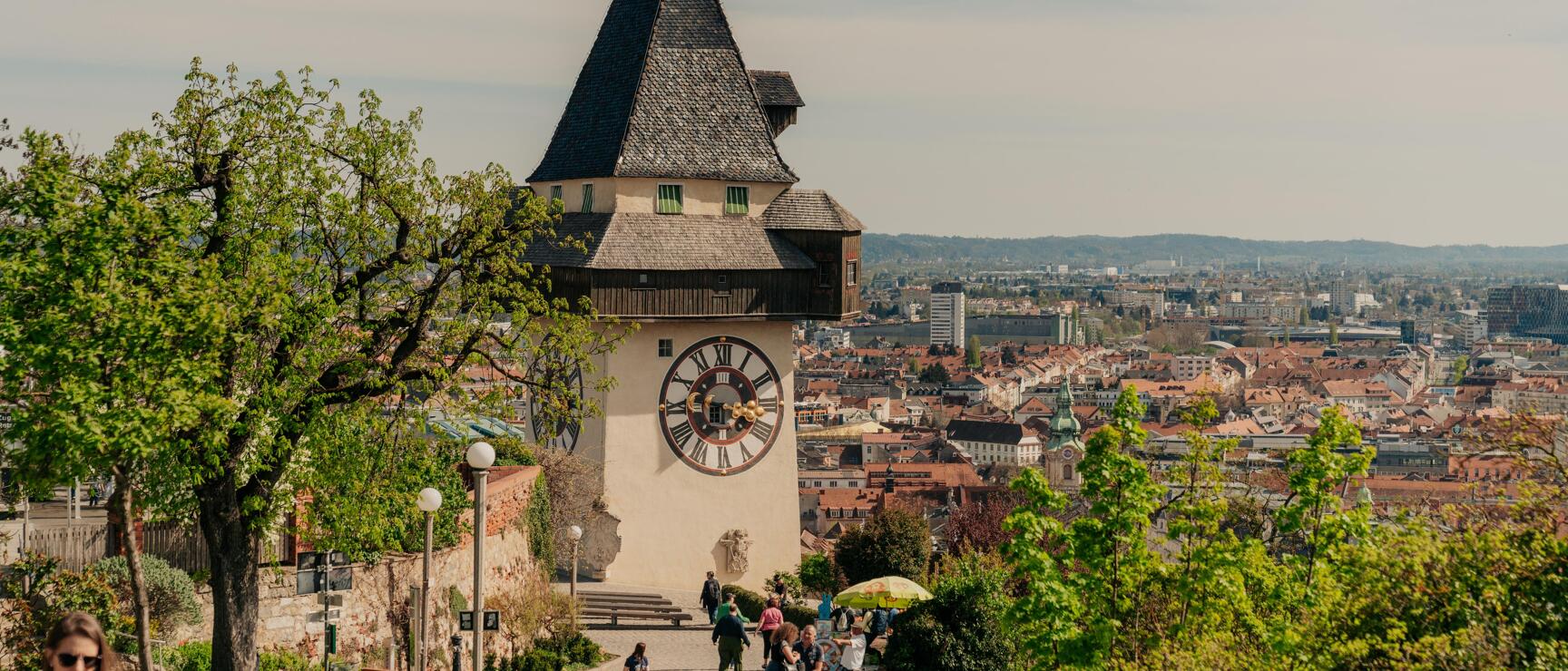 Uhrturm auf dem Schlossberg in Graz mit Blick über die Dächer der Stadt, Menschen auf dem Weg zum Turm.
