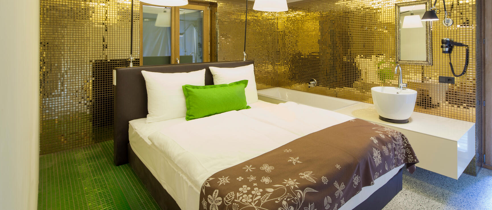 Hotel room with double bed, golden mosaic tiles, green pillow, bathtub and washbasin in the room.