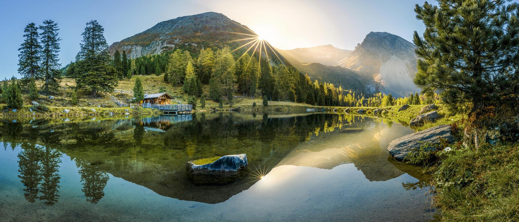 Mirror-calm mountain lake with wooden hut on shore, sun star over mountain peak, coniferous trees, clear sky.
