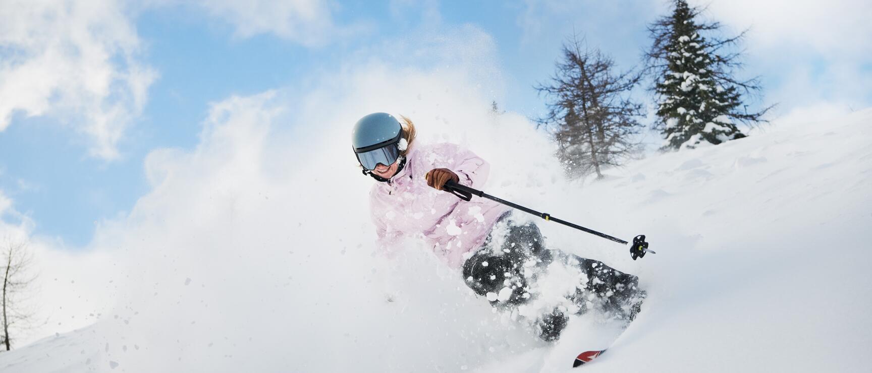 Skier in pink jacket and blue helmet descending through deep powder snow, powder plume, trees in background.