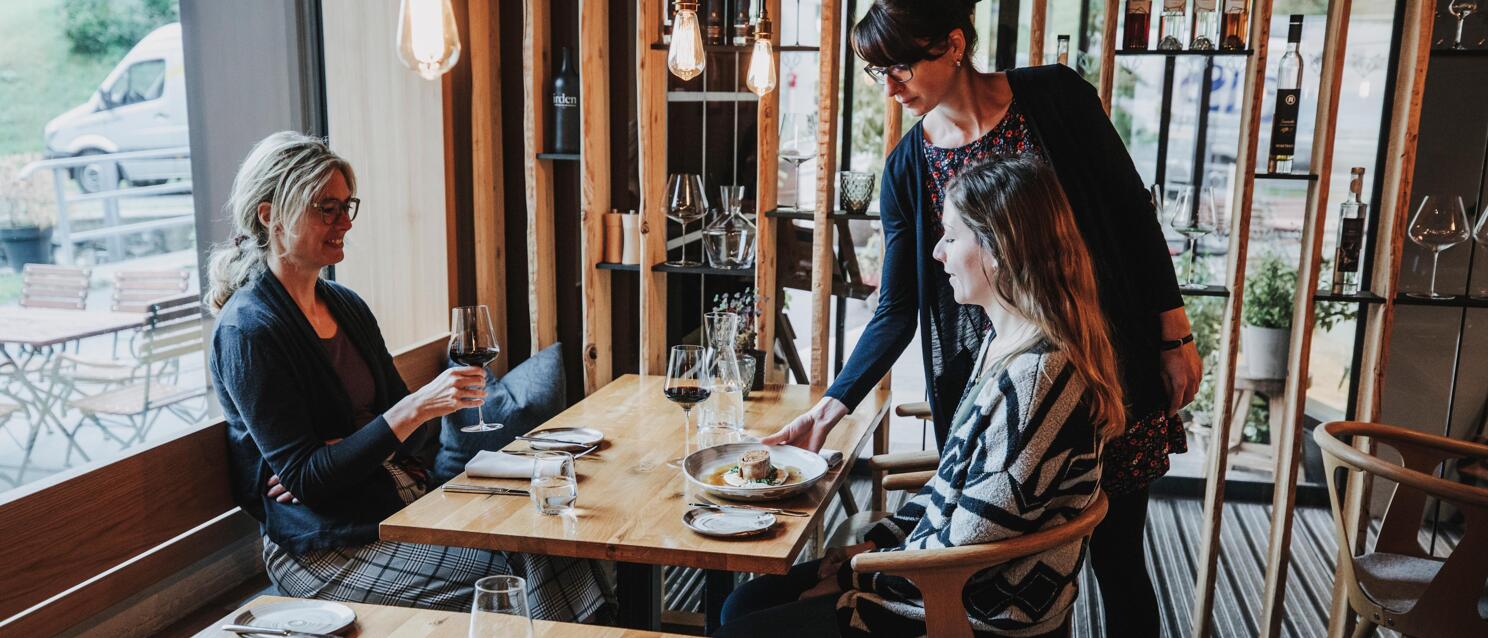 Kellnerin serviert zwei Frauen ein Gericht in einem modernen Restaurant mit Holzmöbeln und Weinregal.