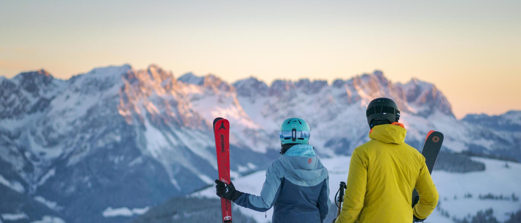 Two skiers holding skis stand on a mountain peak gazing at snow-covered summits in evening light.