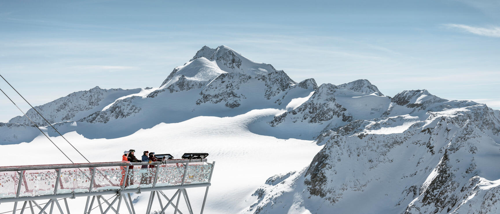 Three people on a metal observation platform overlooking a snowy glacier panorama with mountain peaks.