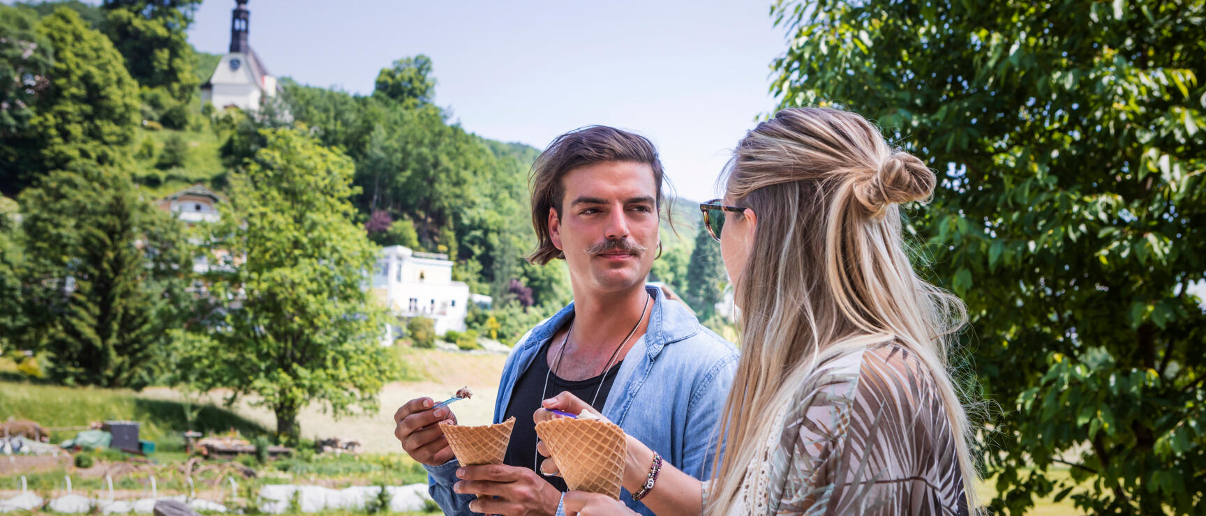 Two people eating ice cream cones in a park with a church tower and green hill in the background.