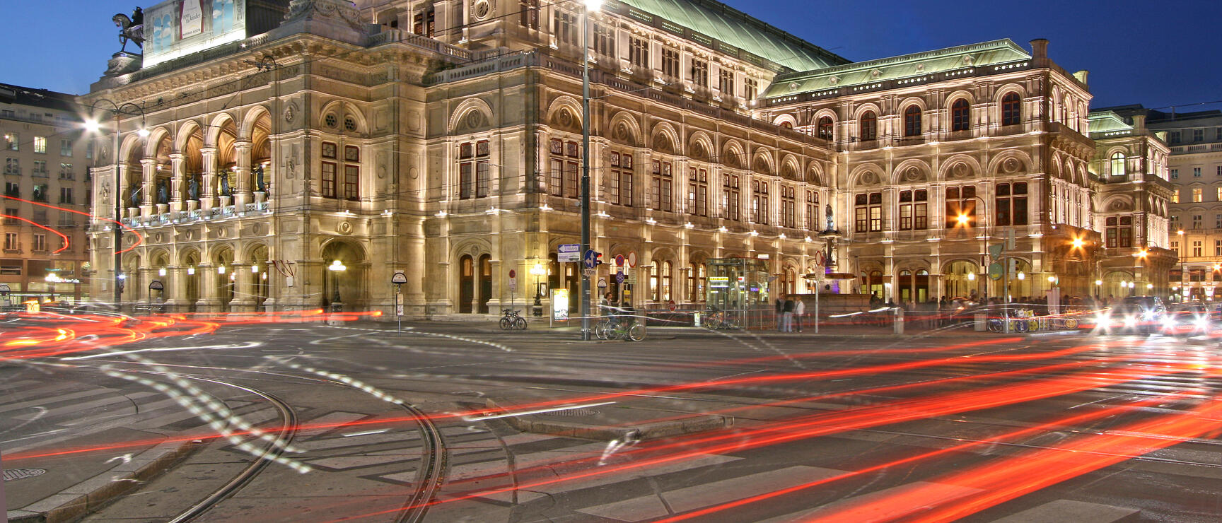 Vienna State Opera at night, illuminated, with red light trails from passing cars in the foreground.