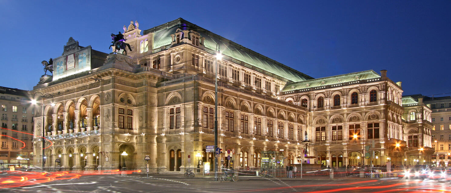 Vienna State Opera at night, illuminated, with red light trails from passing cars in the foreground.