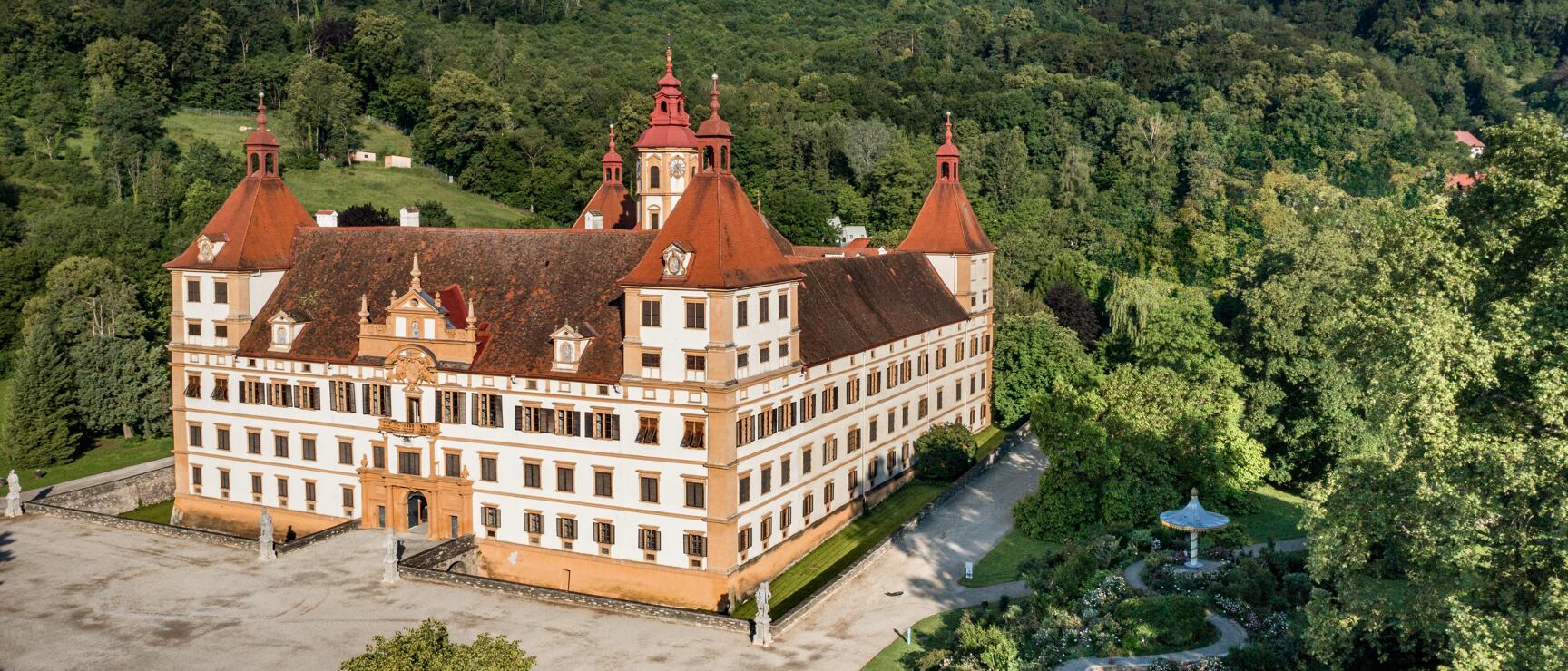 Aerial view of Eggenberg Castle in Graz, white Baroque palace with red towers, park grounds and forested hills.