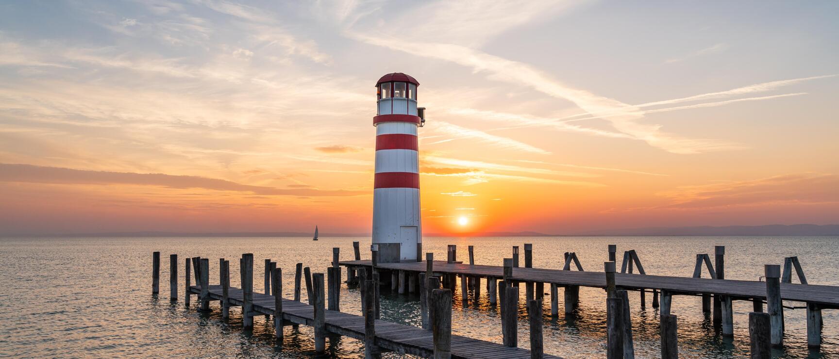 Rot-weiß gestreifter Leuchtturm am Ende eines Holzstegs über dem Neusiedler See bei Sonnenuntergang, mit einem Segelboot am Horizont und orangefarben leuchtendem Himmel.
