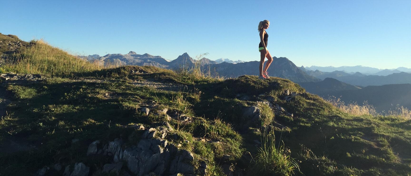 Woman in sports clothing stands on a mountain summit, wide mountain range in background, blue sky.