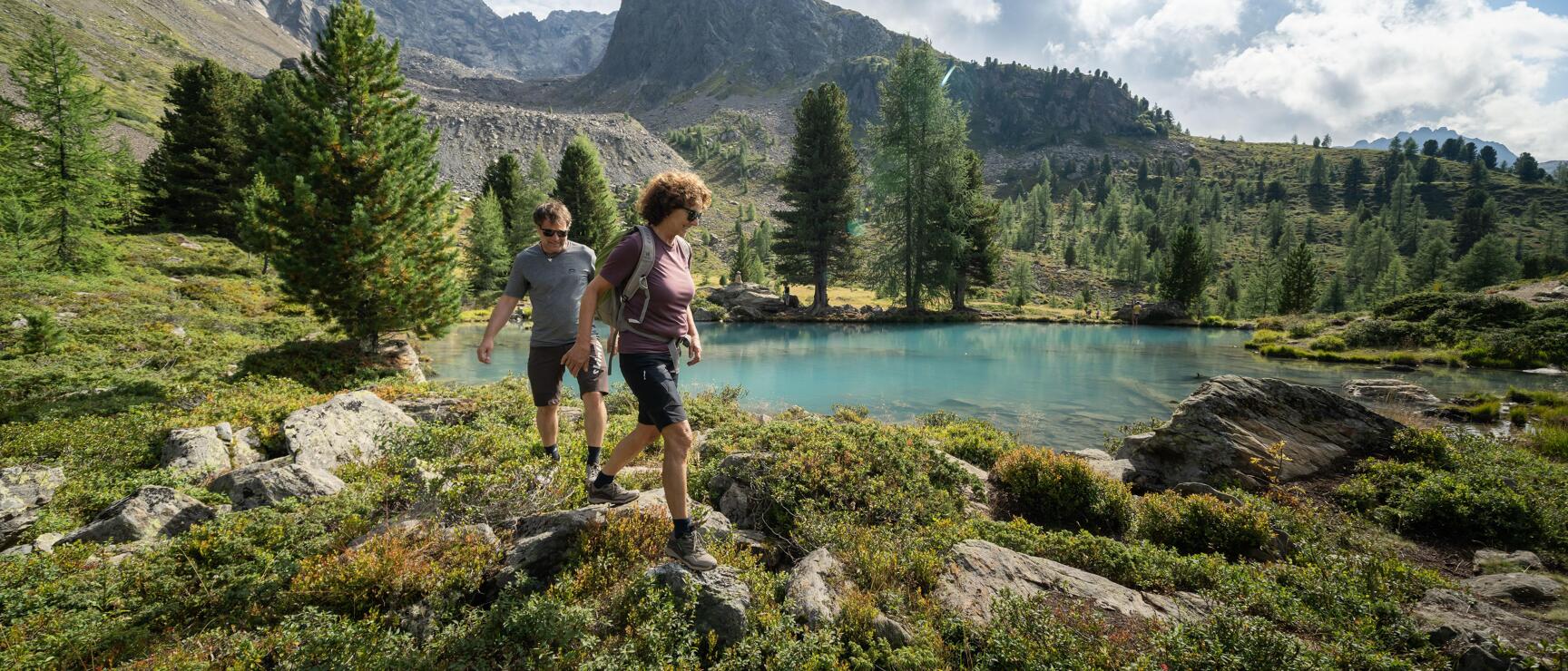 Two hikers on rocky path beside turquoise mountain lake, larch trees and rock peaks in background.
