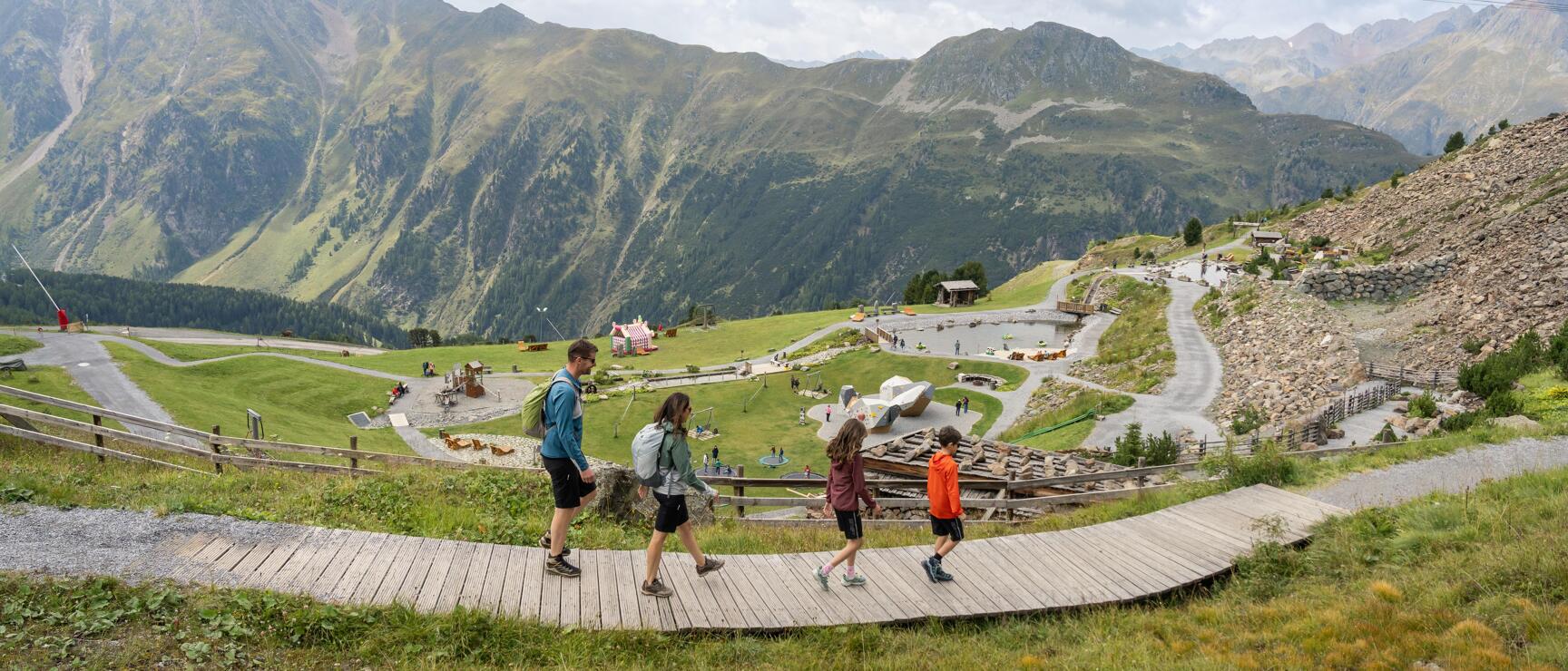 Four people on a wooden jetty in the mountains, cable car and leisure facility in the background.