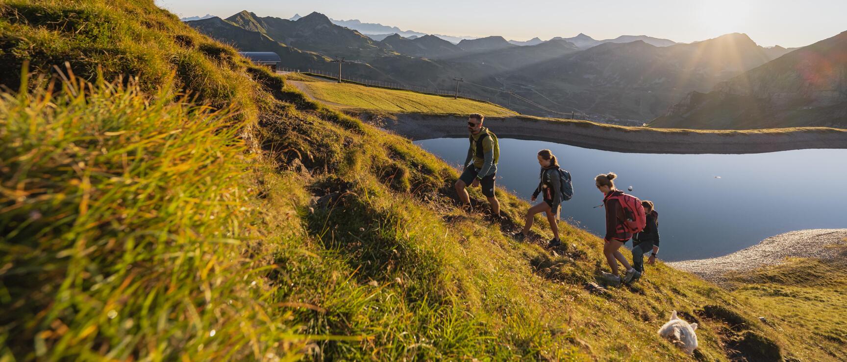 Three hikers with backpacks on a mountain trail next to a reservoir, alpine panorama backlit.