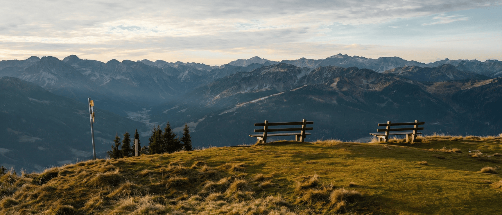 Two wooden benches on an alpine meadow with panoramic mountain range view, overcast sky.