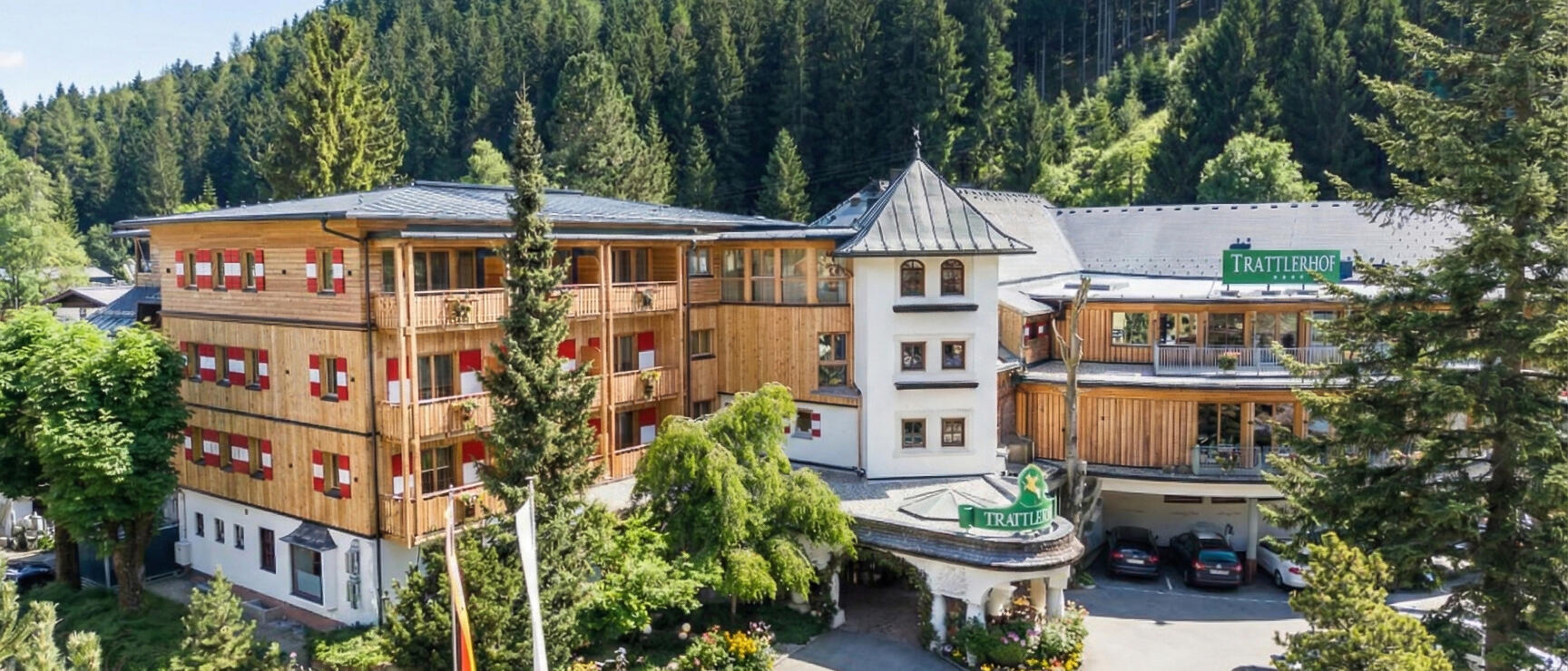 Aerial view of Hotel Trattlerhof with wooden facade, flower beds and dense coniferous forest in background.
