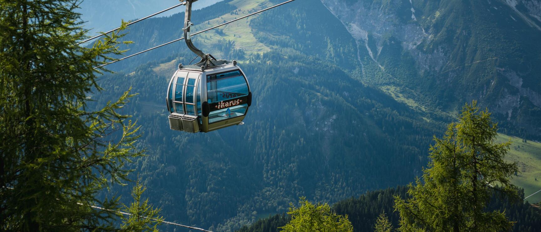 Cable car labeled 'Ikarus' on aerial tramway, surrounded by larch trees, rocky mountain backdrop.