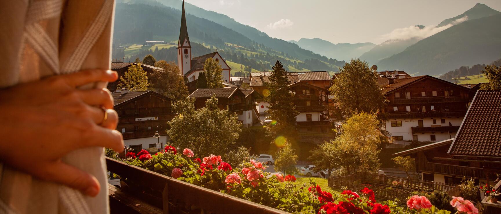 View from balcony with red geraniums over an Alpine village with church tower and mountains in sunlight.