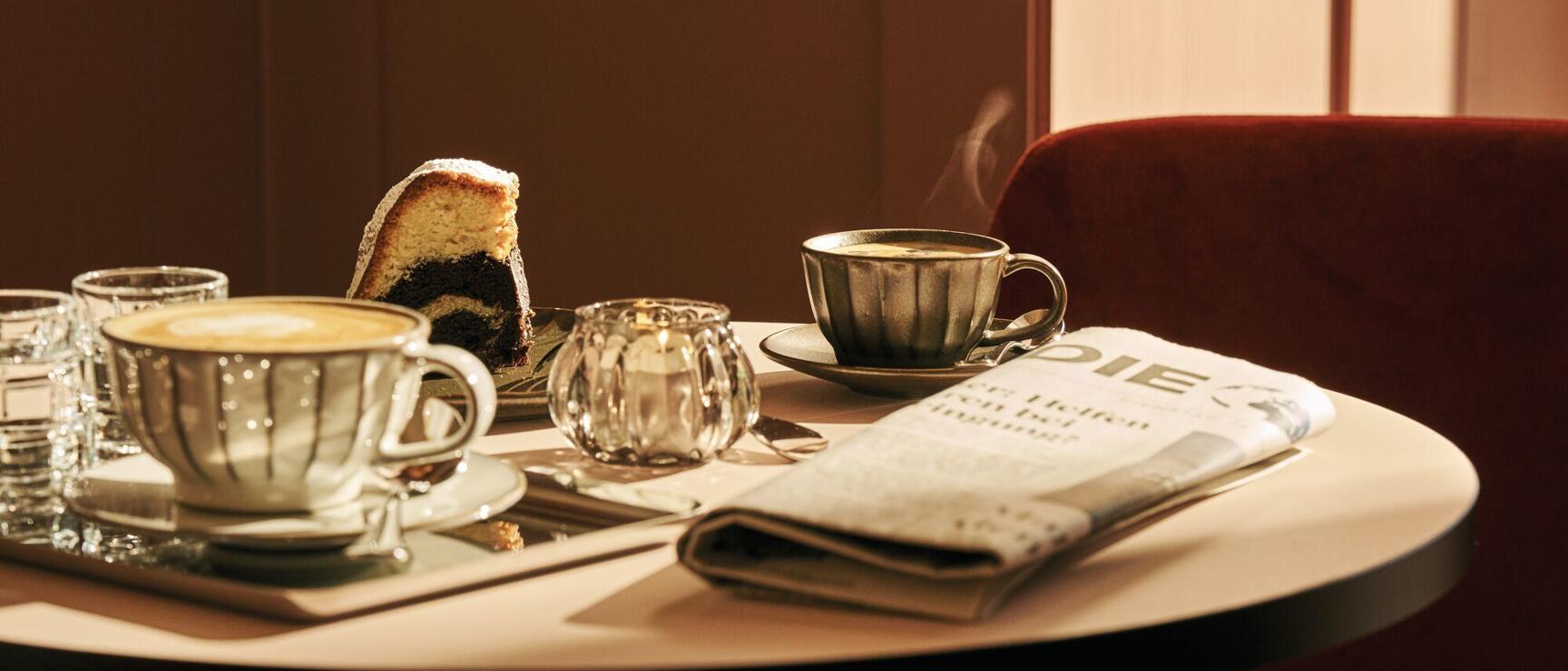 Café house table with cappuccino, steaming coffee, marble cake, candle and newspaper, red velvet armchair in background.
