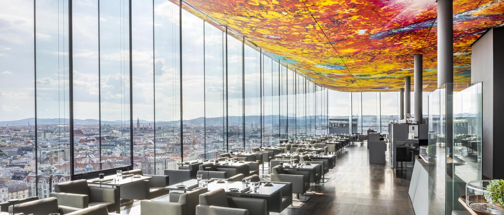 Hochhaus-Restaurant mit bunter Glasdecke, grauen Sesseln und Panoramablick über Wien.