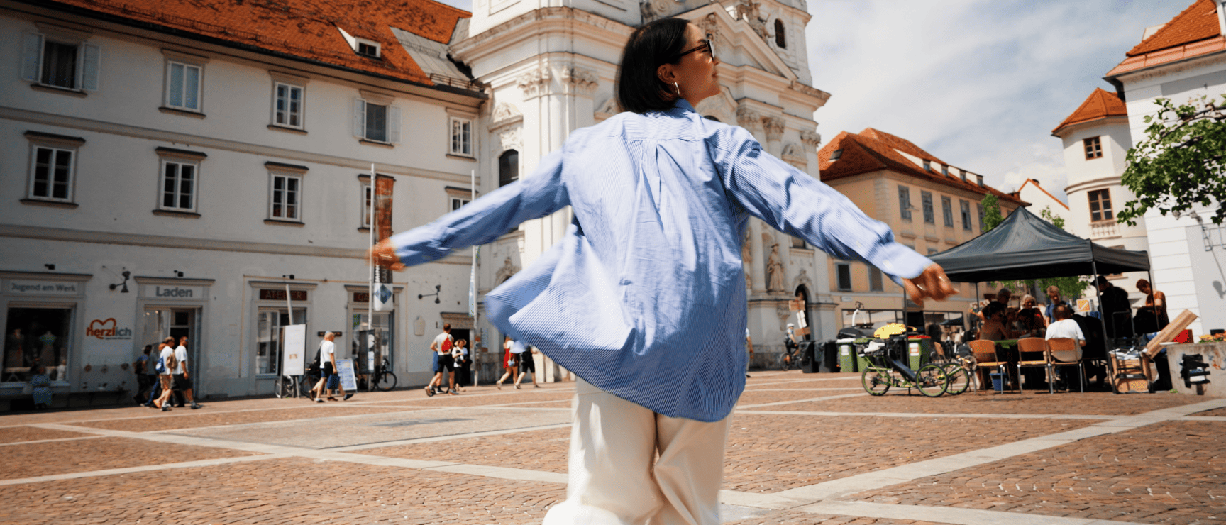 Frau mit ausgebreiteten Armen auf einem Stadtplatz vor einem barocken Kirchturm, Graz.