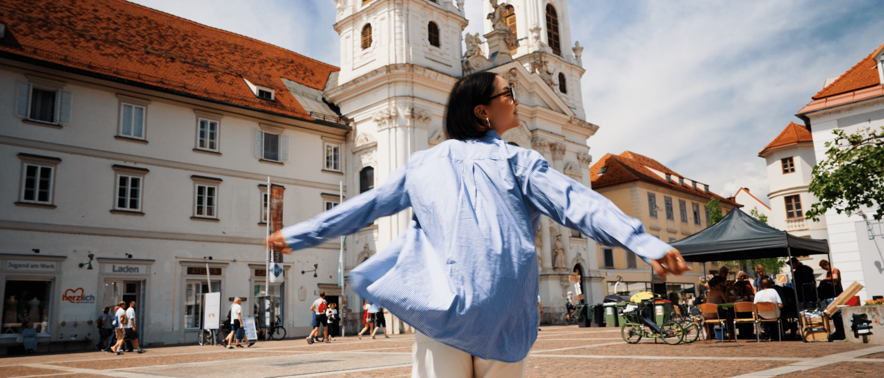 Woman with outstretched arms on a city square in front of a baroque church tower, Graz.