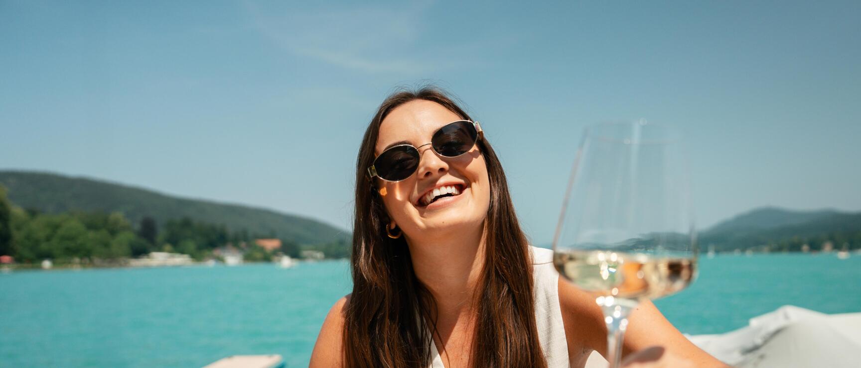 Woman wearing sunglasses holding a wine glass at a lakeside terrace table, turquoise lake and hills in the background.