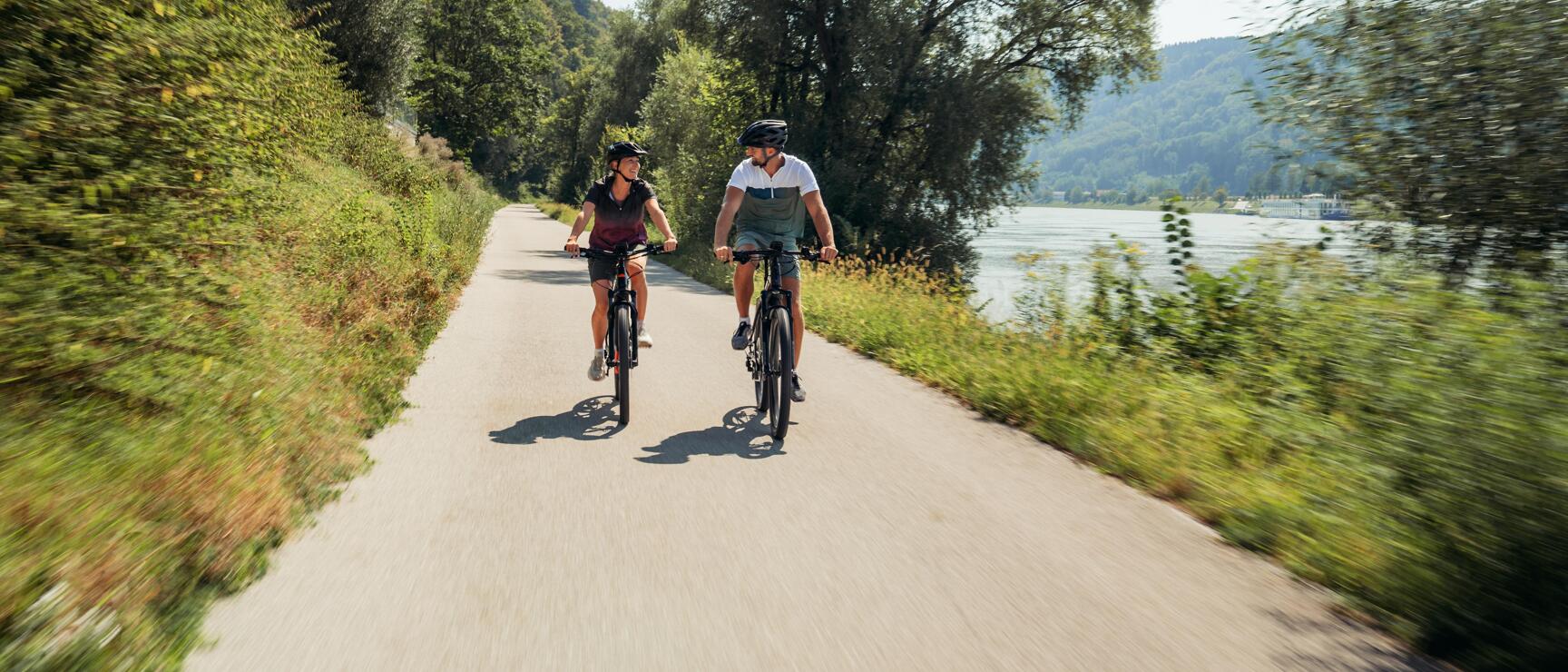 Two cyclists wearing helmets on a path along the Danube with forested hills in the background.