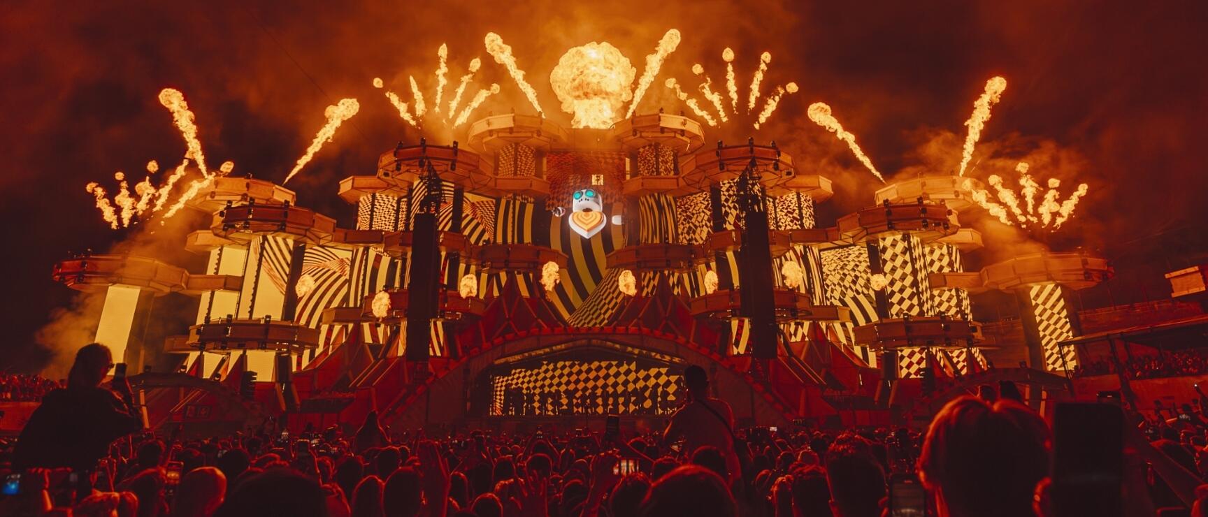Large festival stage with flame effects, fireworks and crowd at night, red light glow.