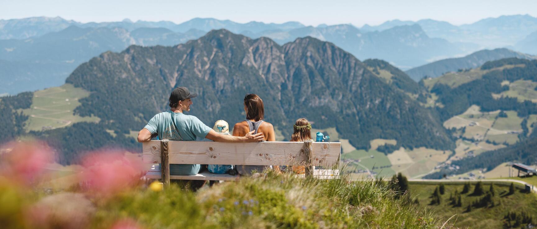 A family sits on a bench on the panoramic trail on the Wiedersbergerhorn and looks out over the surrounding mountains, surrounded by pink alpine roses