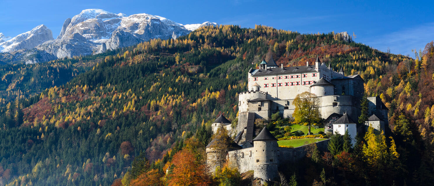 Burg Hohenwerfen auf einem Felsen im Herbst, bunter Wald, schneebedeckte Berge im Hintergrund.