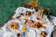 Easter breakfast on a wooden jetty: braided yeast loaves, colourful Easter eggs, ham, tea and juice over turquoise water.