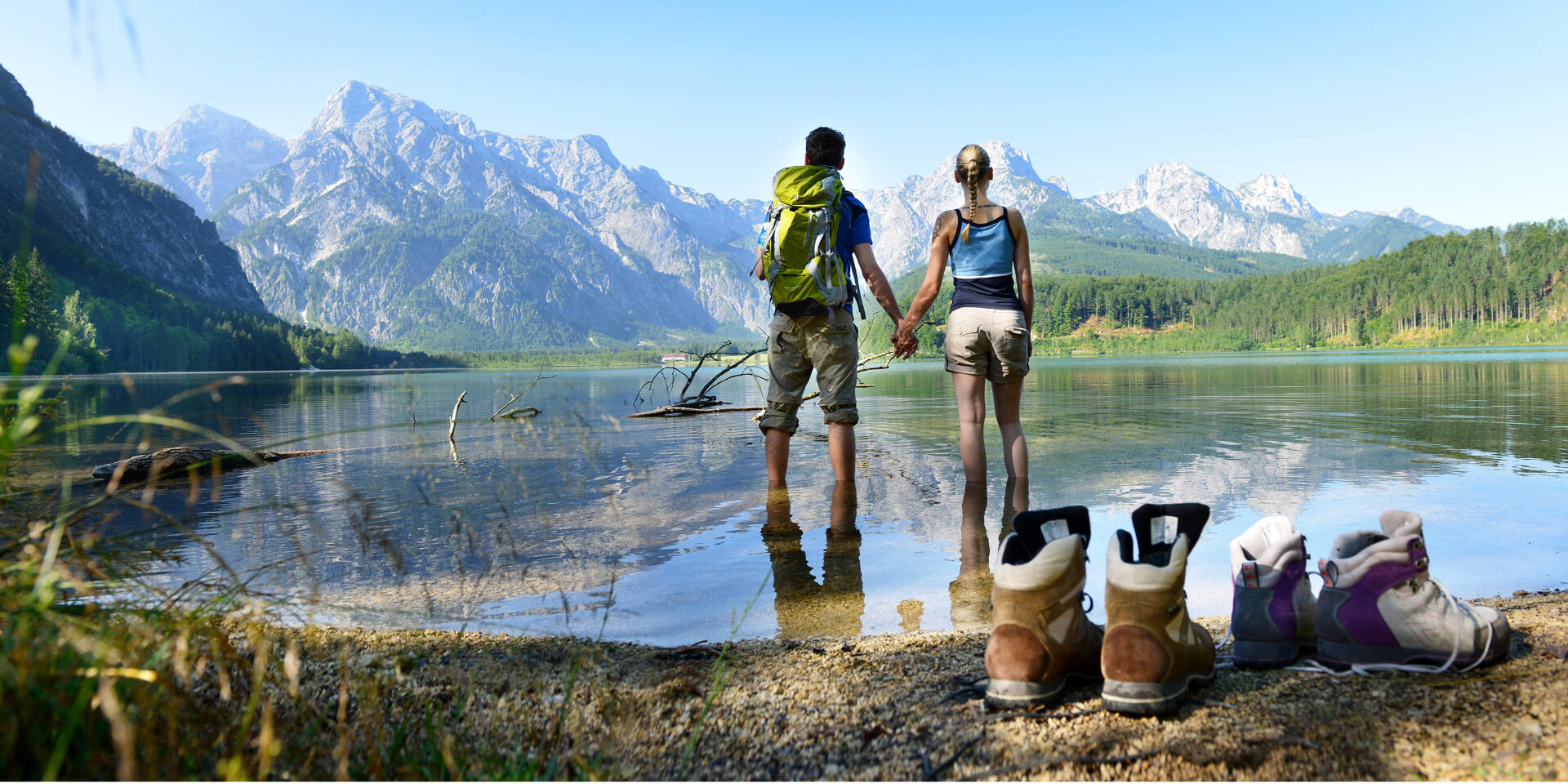 Pure pleasure at the Alm River © Röbl Two people standing barefoot in a mountain lake with hiking boots on the shore and mountain panorama in the background.