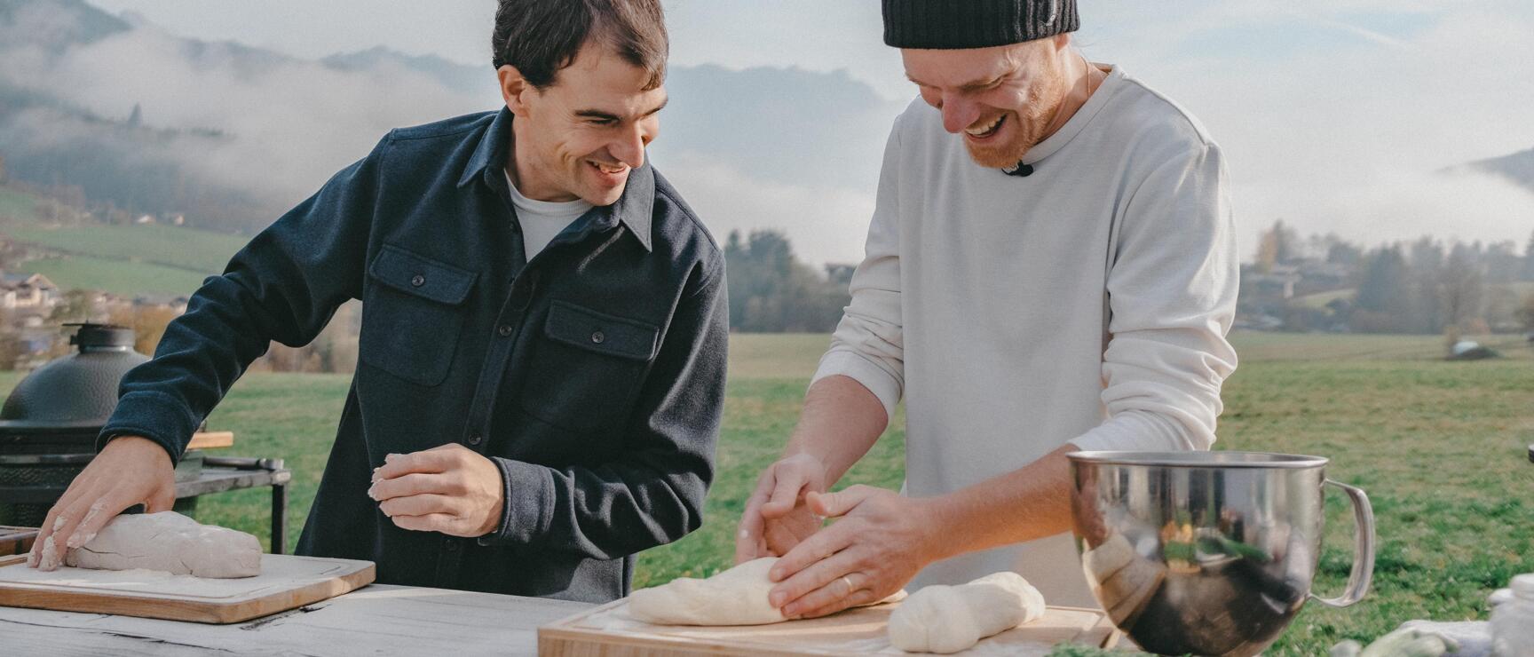 Two men kneading bread dough on wooden boards outdoors, mountainous landscape with meadows in background.
