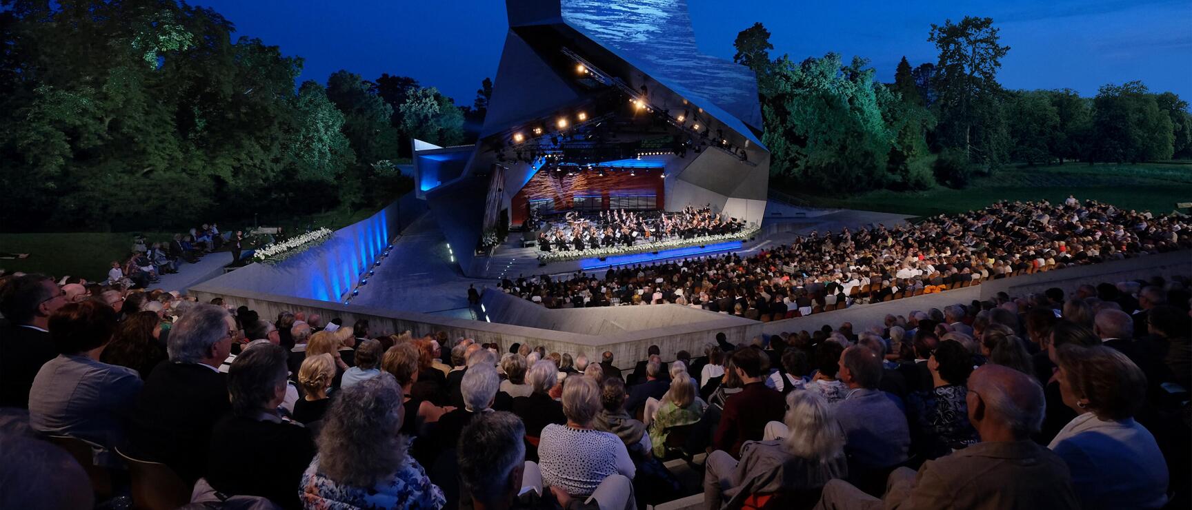 Fully booked open-air amphitheatre at night with orchestra on illuminated stage, modern stage roof, trees in background.