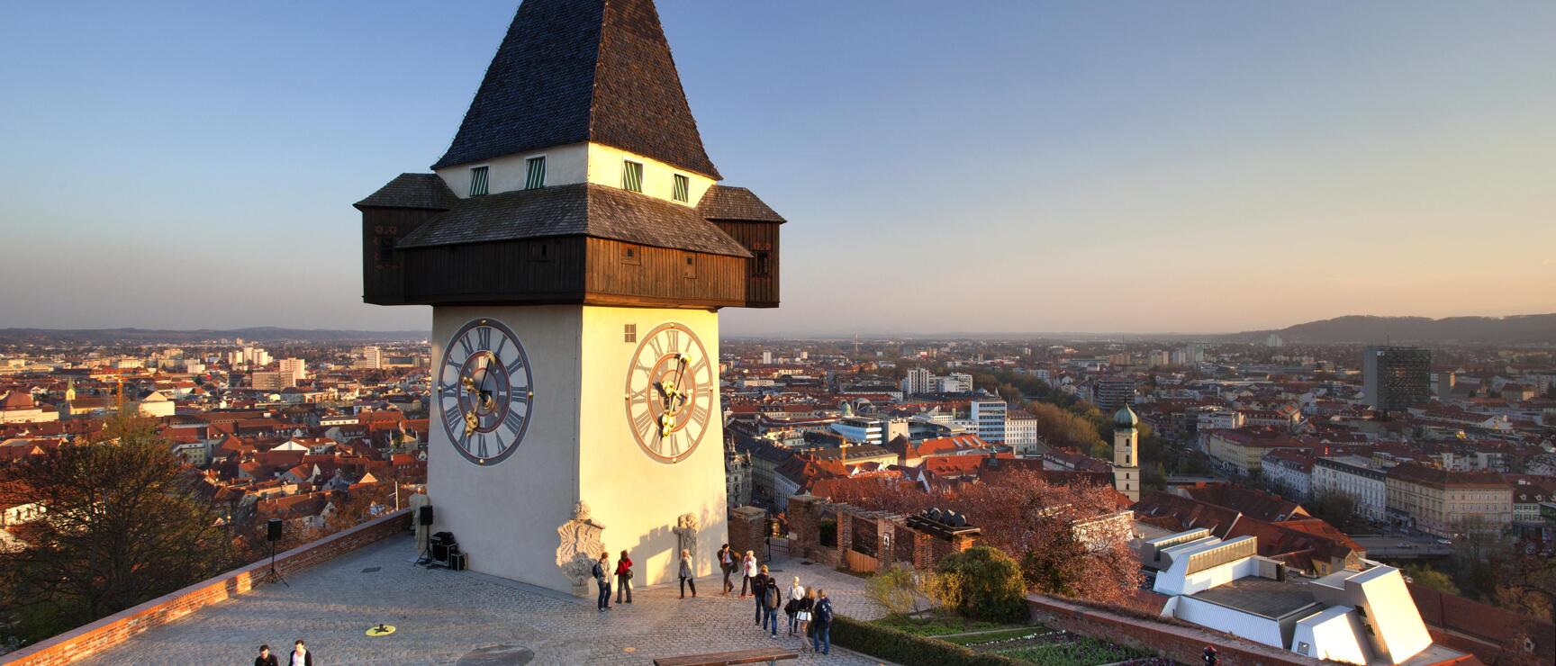 Clock tower on Schlossberg in Graz at sunset light, city panorama in background, visitors on the square.
