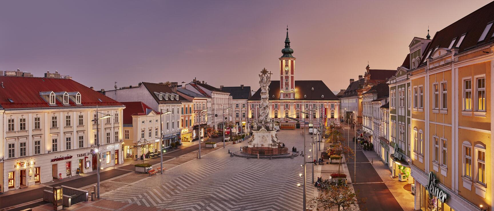 Main square in St. Pölten at dusk with Trinity Column, illuminated town hall and surrounding Baroque facades.