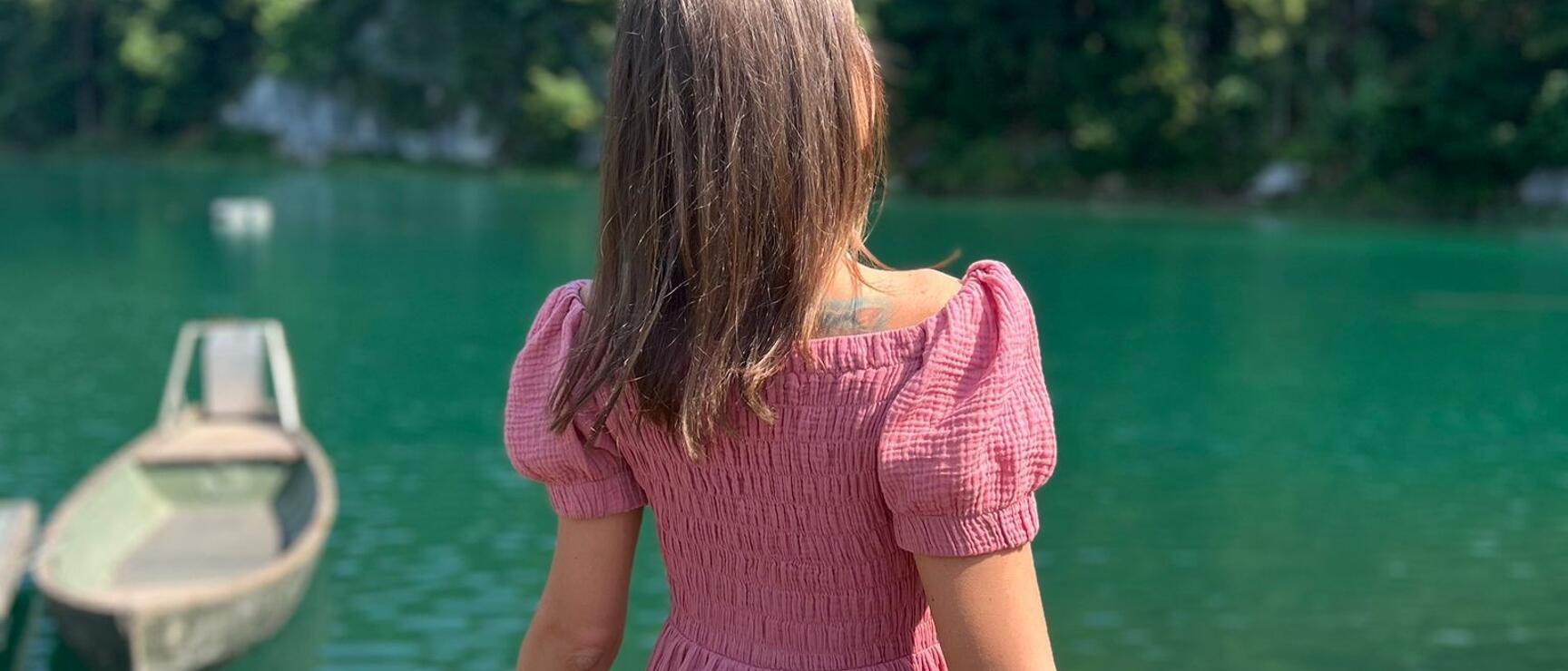 Frau im rosa Kleid blickt auf türkisblauen See mit Ruderboot, bewaldete Hügel im Hintergrund.