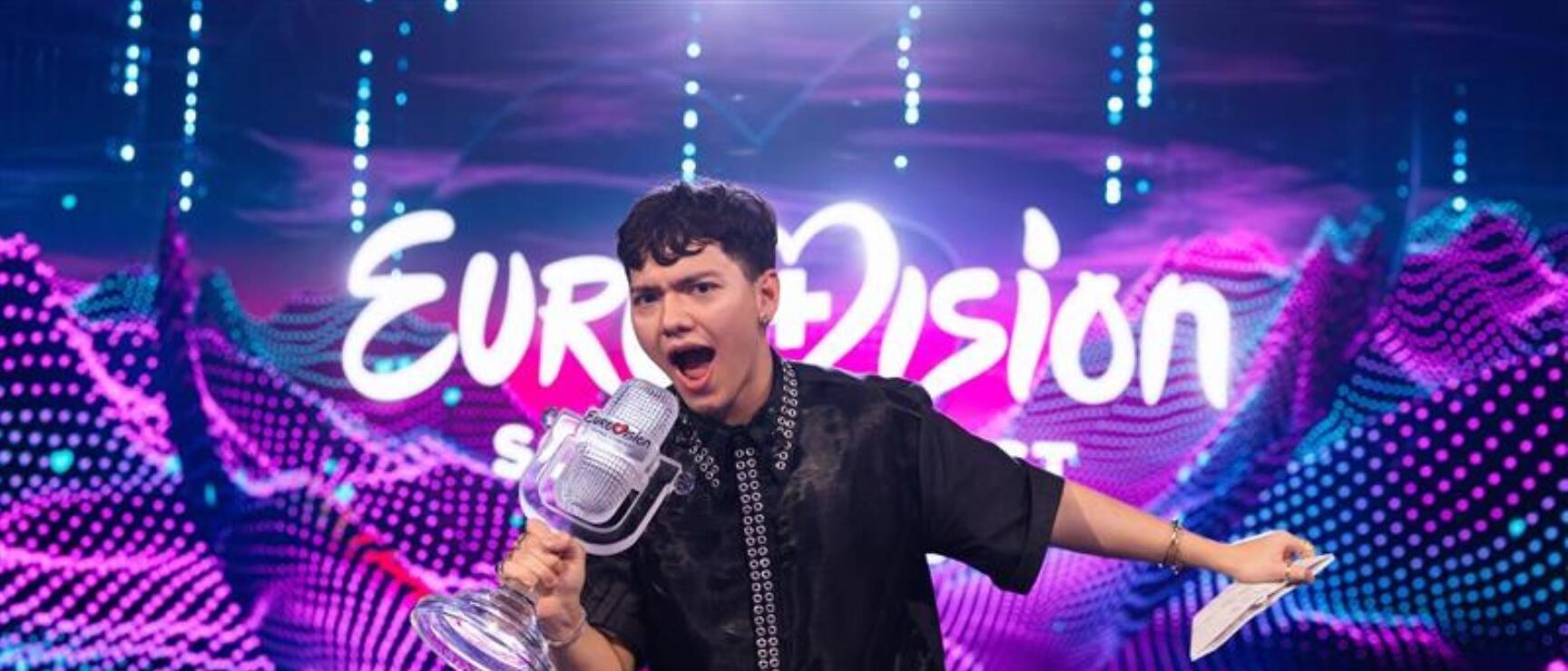 Person in black outfit holding Eurovision trophy in front of brightly coloured Eurovision Song Contest stage.