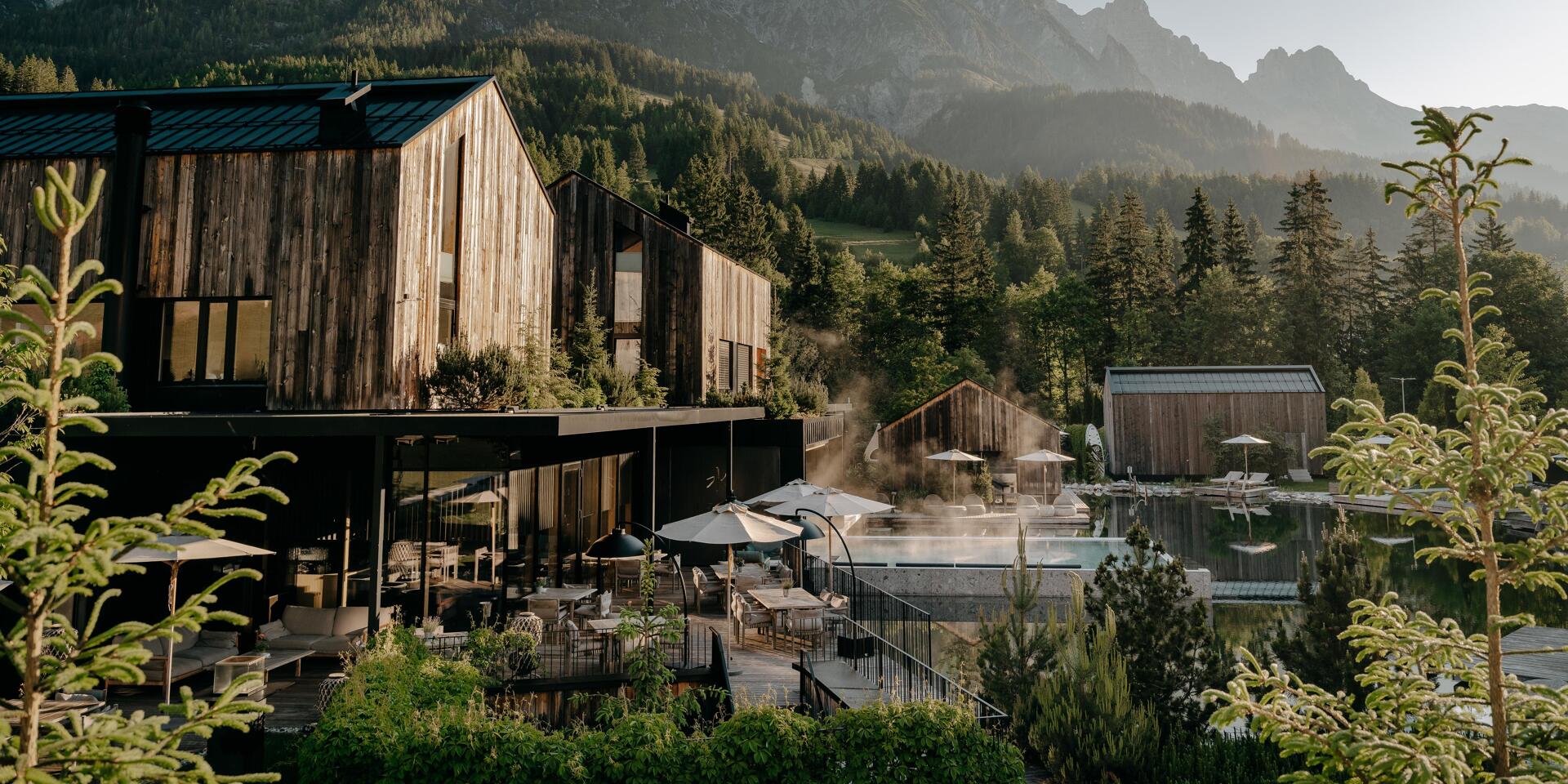 Wooden hotel with steaming outdoor pool, sun umbrellas and terrace, coniferous forest and rocky peak in background.