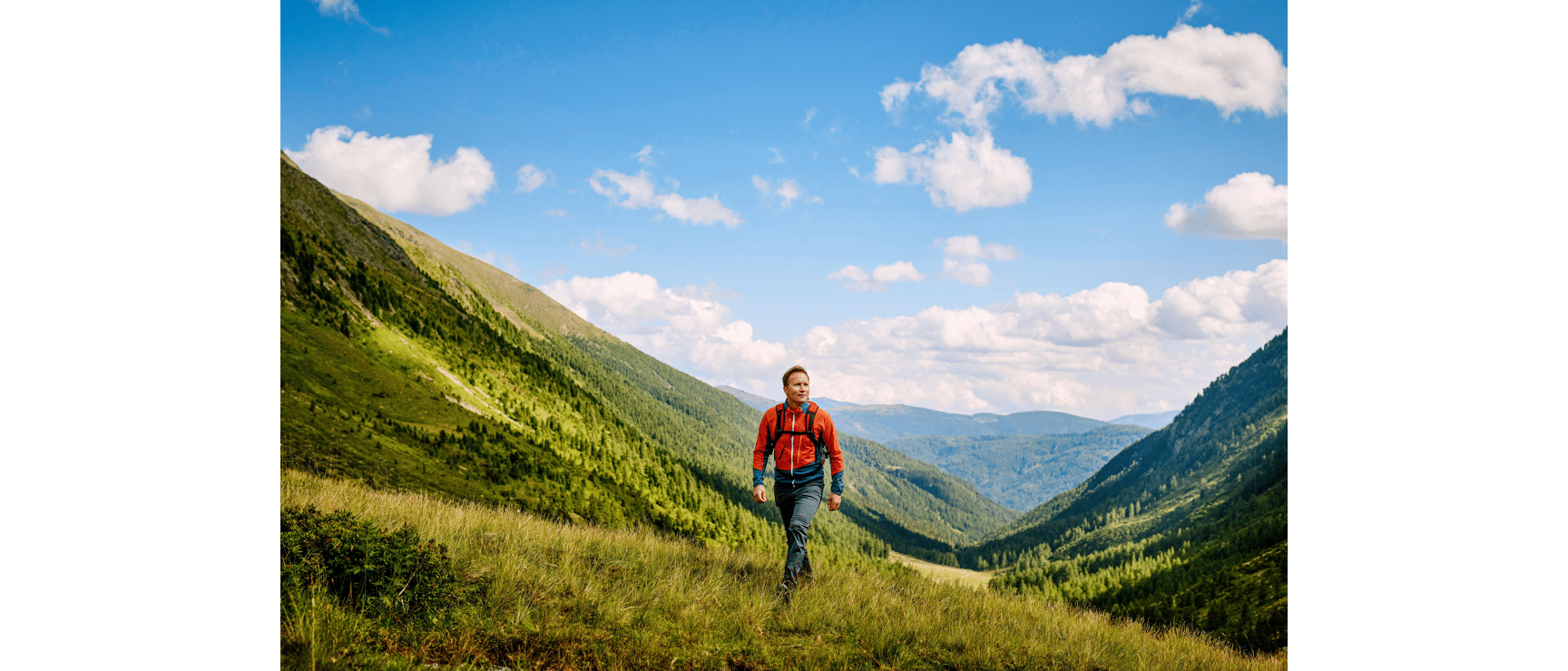 Man in red anorak and backpack hiking across alpine meadow with green mountain valley and blue sky in background.