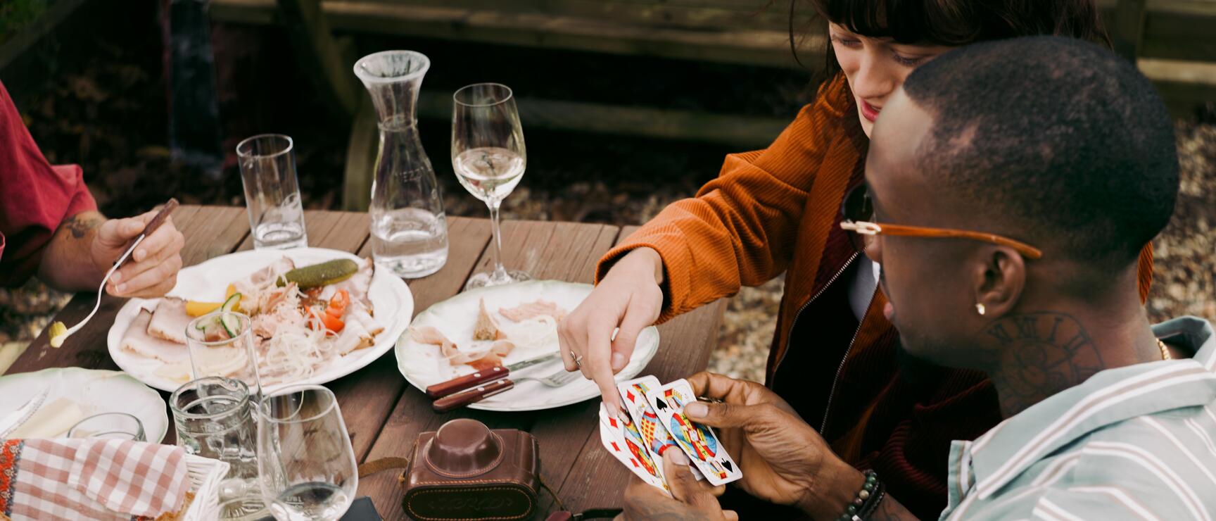 Zwei Personen spielen Karten an einem Holztisch im Freien mit Speisen, Brot und Rotweingläsern.